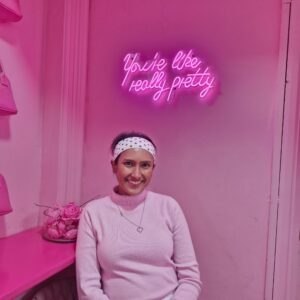 Elizabeth is smiling to the camera, wearing pink clothes and a headband. She is in a pink room with a pink background.