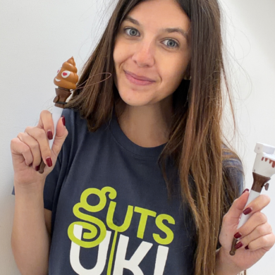 Sophie stands in front of the camera, wearing a navy Guts UK t-shirt and smiles. She has long brown hair and is holding a Guts UK poo emoji pen in each hand.