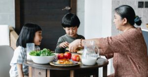 A family sitting at a table eating food. The woman is serving the young boy and girl. They are smiling and talking in their kitchen, sitting down at the table. The table has healthy food like fruit and vegetables. There are also jugs of milk on the table.