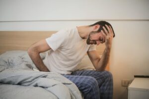 A man in bed wearing pyjamas sitting on a bed with his hand in his head looking tired and in discomfort 