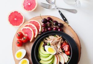 A picture of healthy food, the meal includes eggs, fruits, fish and vegetables on a table, with a knife and fork next to it.